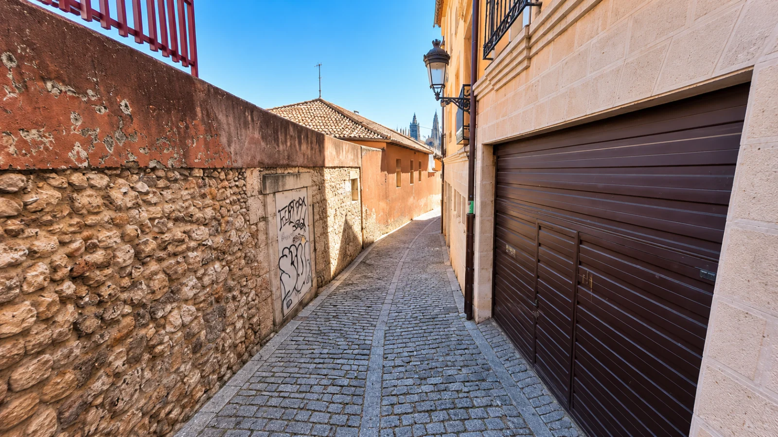 Vista del callejón empedrado delimitado por un muro de mampostería con las agujas de la Catedral asomando al fondo