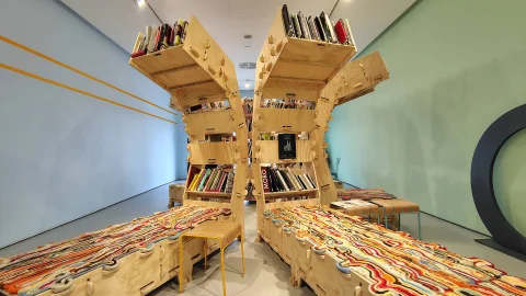 Perspectiva frontal de dos estructuras de madera que combinan estantes para libros y tumbonas con tapicería artesanal en coloridos patrones