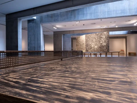 Primer plano de una mesa de ping-pong de madera con red metálica en una sala del museo con vigas de hormigón y murales al fondo
