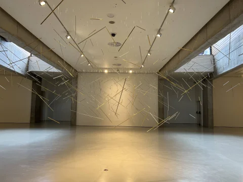 Sala de exposición diáfana con decenas de varillas metálicas doradas colgadas del techo en una composición flotante