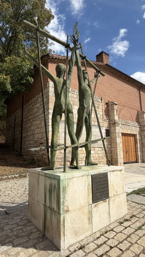 Escultura de bronce con figuras humanas y rejas frente a la fachada de ladrillo y piedra del Teatro Clunia