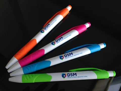 Cuatro bolígrafos naranja, rosa, azul y verde con el logotipo de DSM sobre una superficie negra